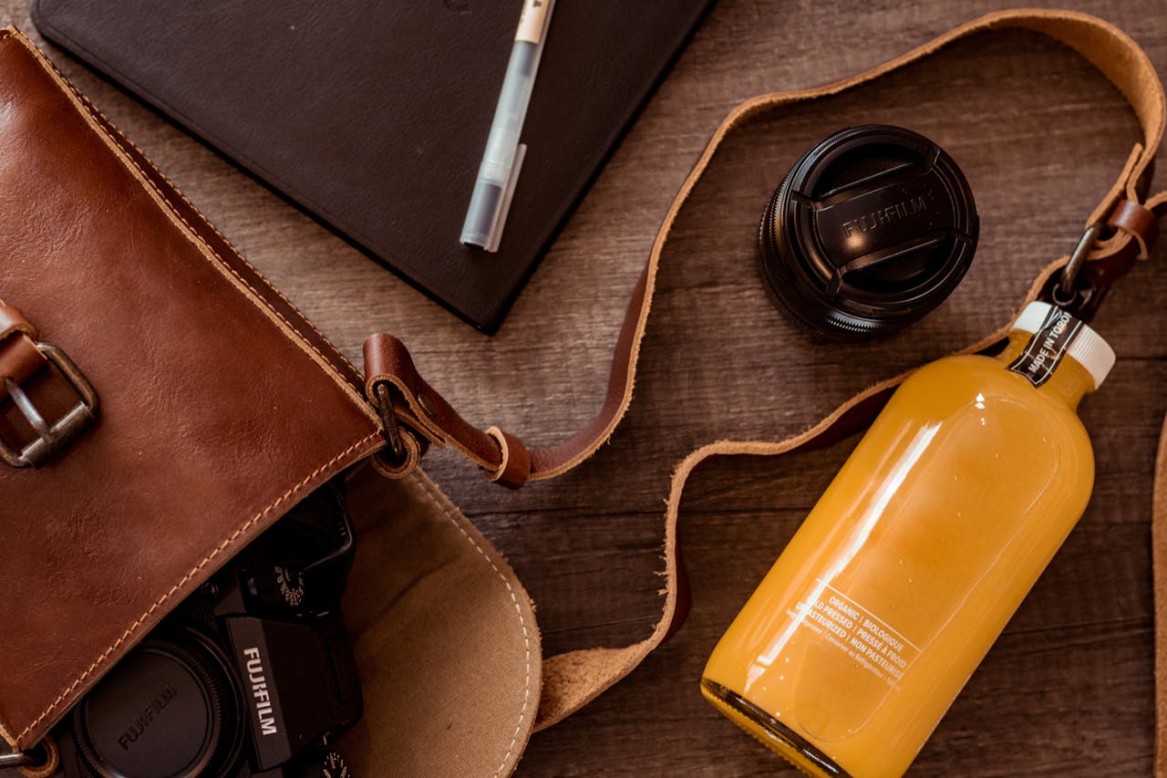 Stylish flat lay of leather bag, camera lens, and refreshing orange beverage on wooden surface.